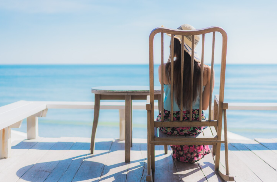 Woman wearing a sun hat sitting on a wooden chair by the ocean, enjoying a budget-friendly beach vacation